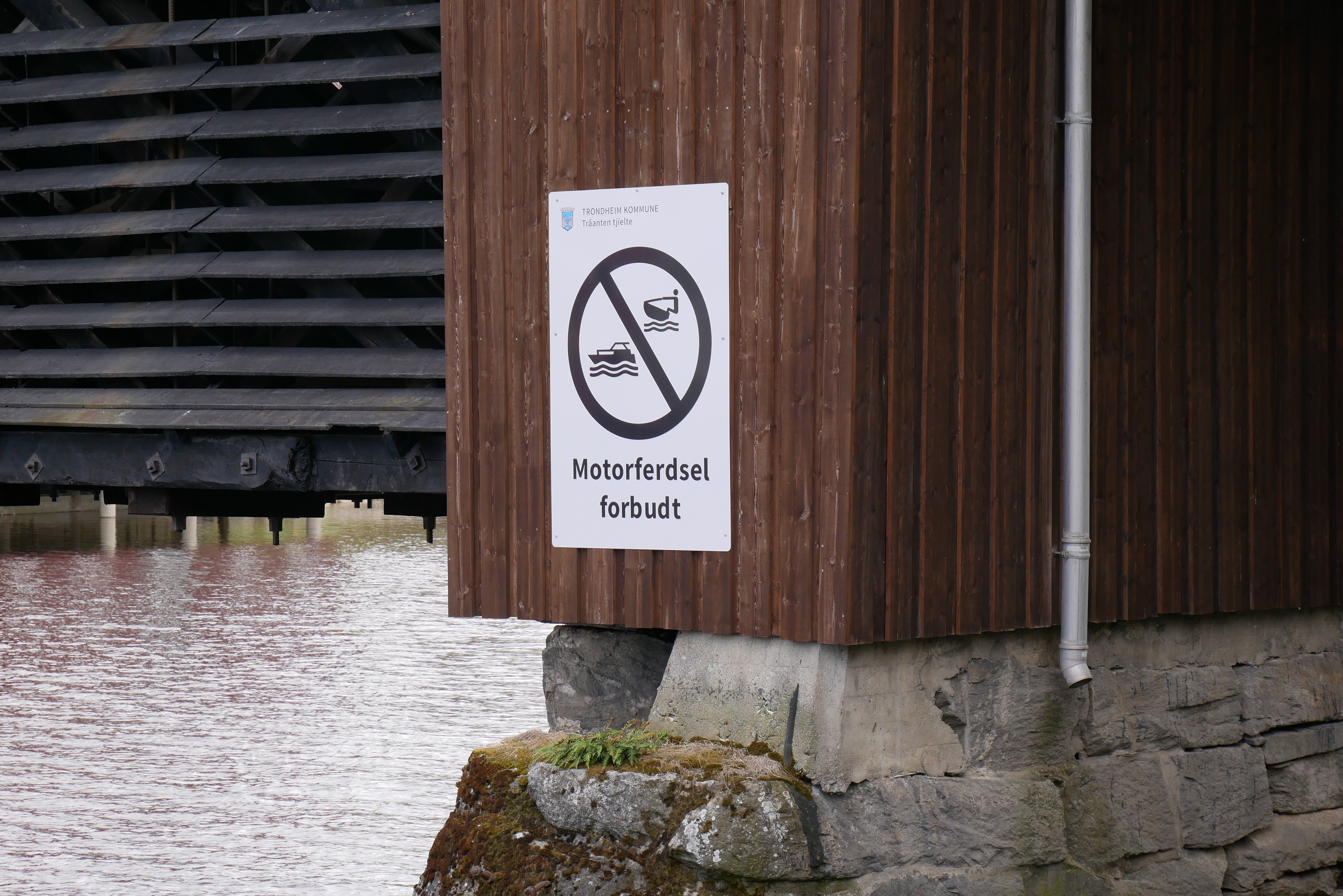 Et hvitt skilt med et forbudssymbol som viser en motorb&aring;t og teksten &laquo;Motorferdsel forbudt&raquo; er festet til en brun trekonstruksjon, sannsynligvis en bro, over en elv.