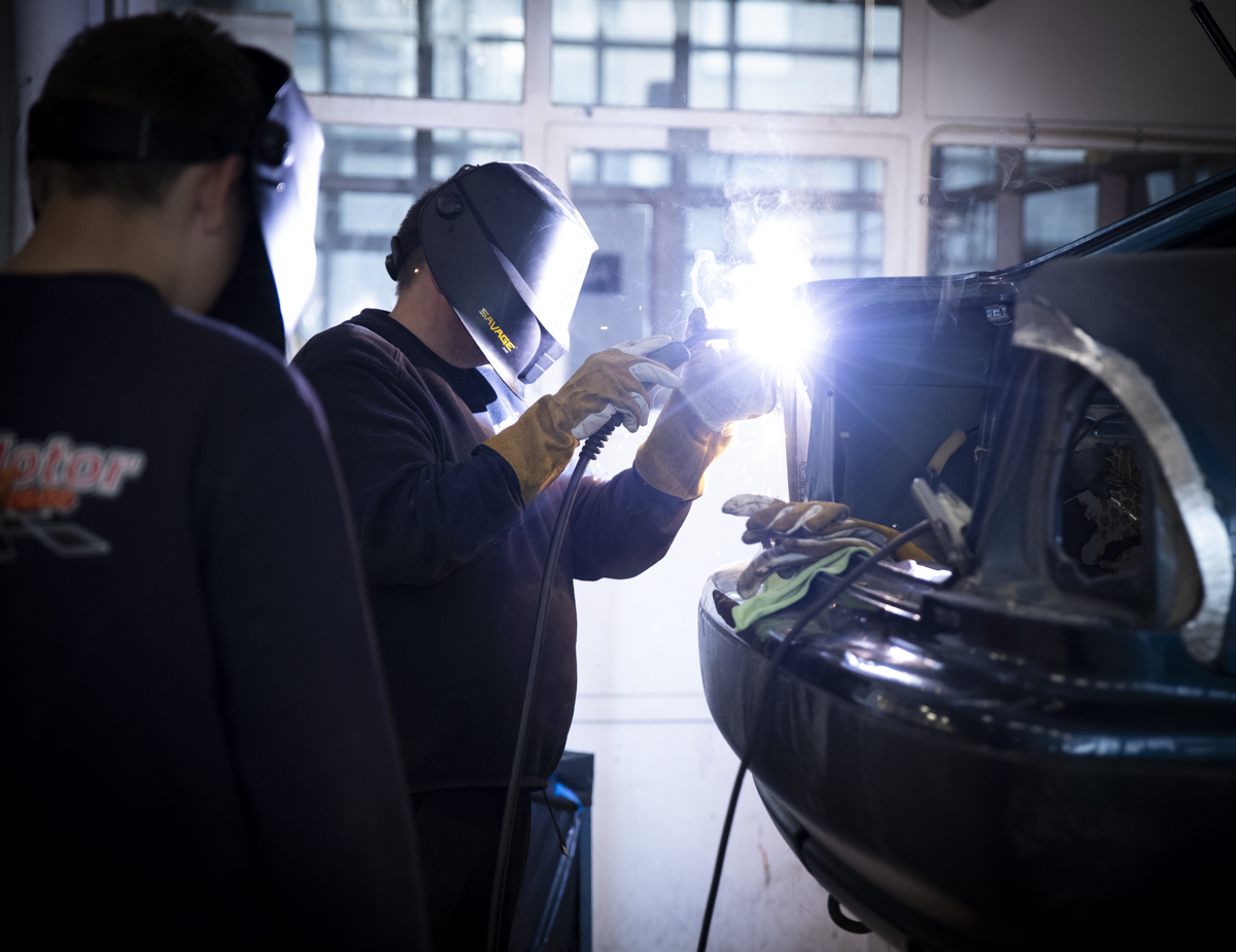 En person sveiser på en bil, mens en annen står og ser på. 