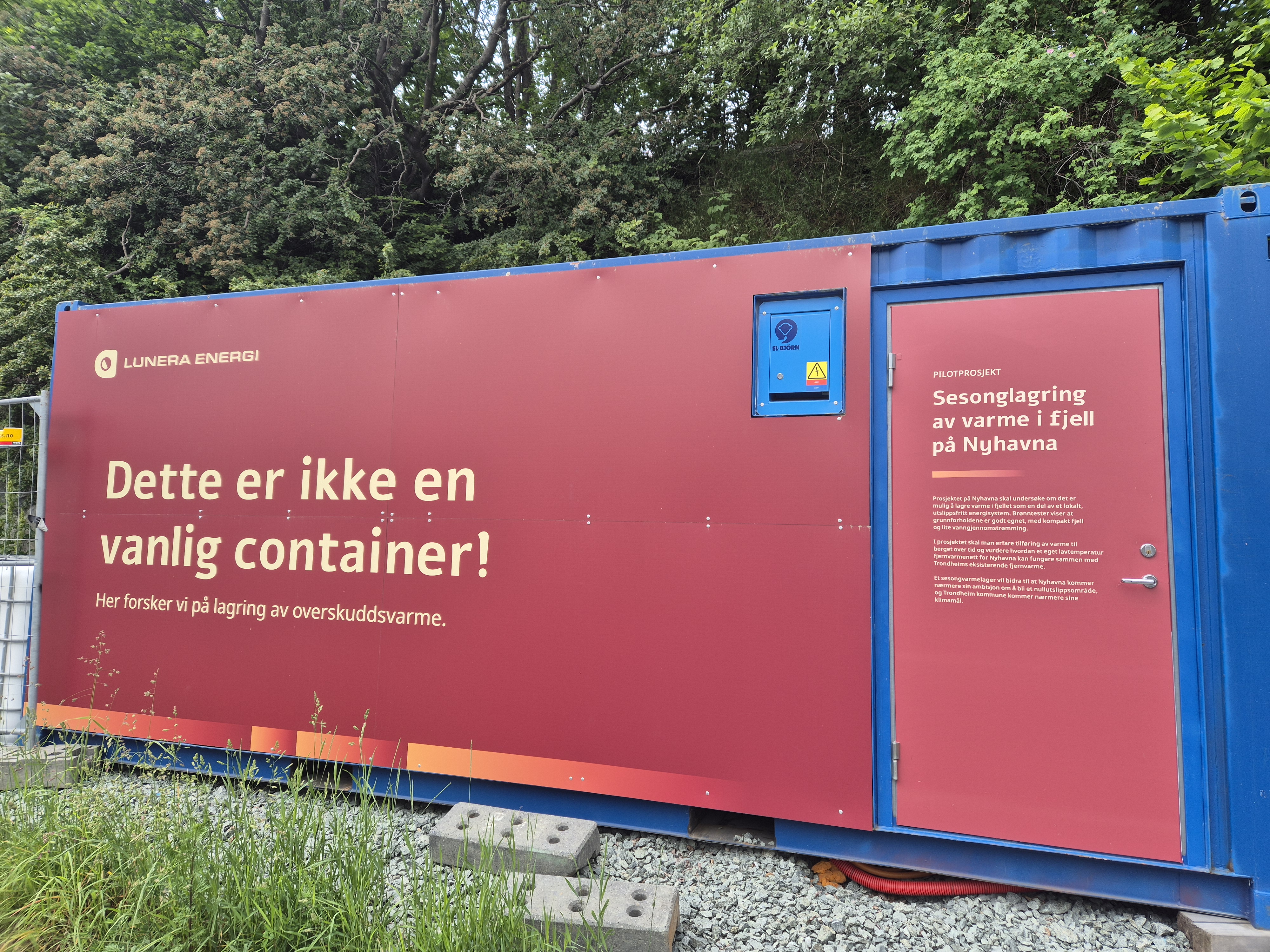 En stor, bl&aring; container med r&oslash;d kledning st&aring;r plassert utend&oslash;rs foran gr&oslash;nne tr&aelig;r, dekket med hvit og gul tekst fra Lunera Energi om et pilotprosjekt for sesonglagring av overskuddsvarme i fjell p&aring; Nyhavna. Teksten inkluderer hovedoverskriften &laquo;Dette er ikke en vanlig container! Her forsker vi p&aring; lagring av overskuddsvarme.&raquo; samt en lengre infotekst p&aring; d&oslash;ren. Infotekst p&aring; d&oslash;ren: LUNERA ENERGI  Dette er ikke en vanlig container! Her forsker vi p&aring; lagring av overskuddsvarme. PILOTPROSJEKT Sesonglagring av varme i fjell p&aring; Nyhavna  Prosjektet p&aring; Nyhavna skal unders&oslash;ke om det er mulig &aring; lagre varme i fjellet som en del av et lokalt, utslippsfritt energisystem. Br&oslash;nntester viser at grunnforholdene er godt egnet, med kompakt fjell og lite vanngjennomstr&oslash;mming.  I prosjektet skal man erfare tilf&oslash;ring av varme til berget over tid og vurdere hvordan et eget lavtemperatur fjernvarmenett for Nyhavna kan fungere sammen med Trondheims eksisterende fjernvarme.  Et sesongvarmelager vil bidra til at Nyhavna kommer n&aelig;rmere sin ambisjon om &aring; bli et nullutslippsomr&aring;de, og Trondheim kommune kommer n&aelig;rmere sine klimam&aring;l.