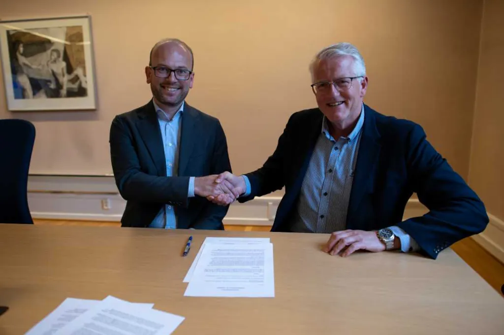 Kristian Dahlberg Hauge og Svein Ove Slinde sitter ved siden av hverandre. De håndhilser mens avtalen de har signert ligger foran dem på bordet. Begge smiler.