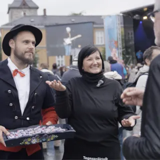 En mann i tradisjonelle kostymer sammen med en kvinnelig tolk, vi ser ryggen til en eldre mann som følger med.