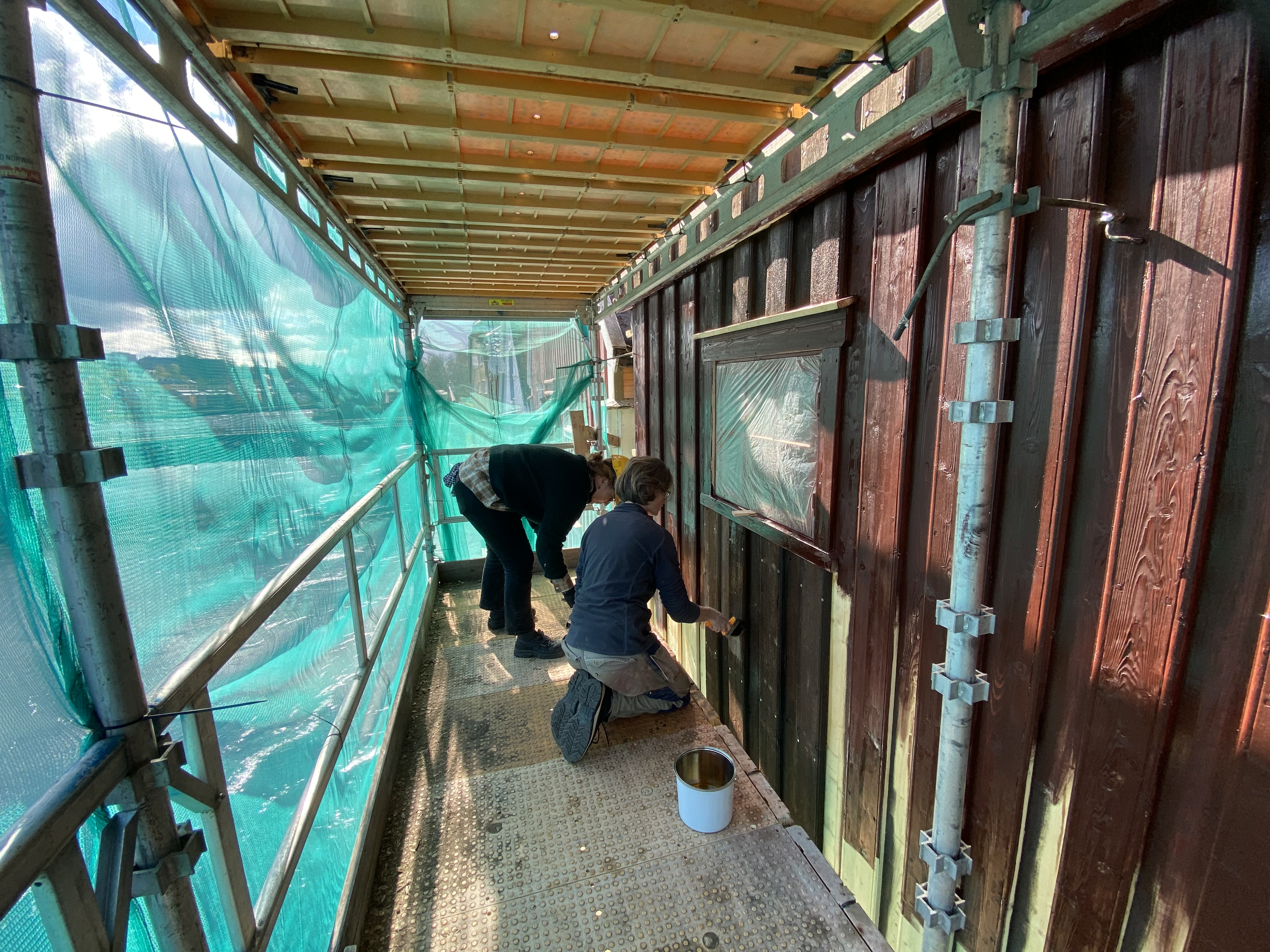 To personer sitter på et stillas og maler en vegg