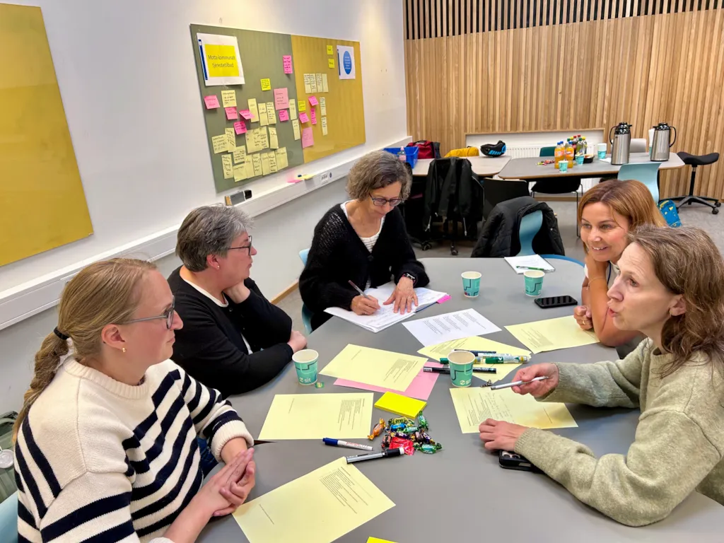 Fem kvinner sitter rundt et bord og jobber med oppgaver i en workshop