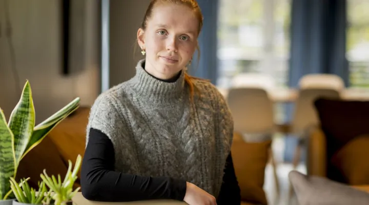 Lærling Thea Udness sitter på en stol i stua på Byåsen BOA. Bakgrunnen er diffus, mens Thea ser i kamera. Vi ser den øvre delen av kroppen hennes. I forkant ser vi planter i venstre hjørne.