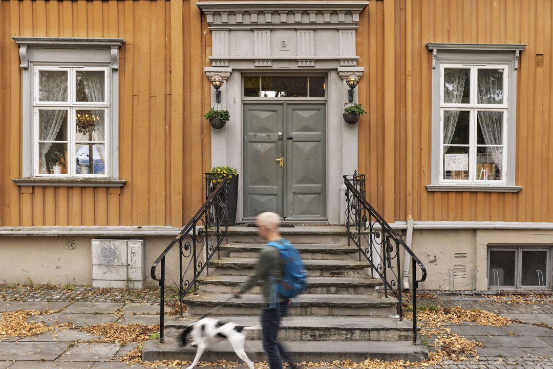 av okergul trebygning med en stor steintrapp utenfor som fører opp til en dobbel inngangsdør. Mann med hund går forbi.