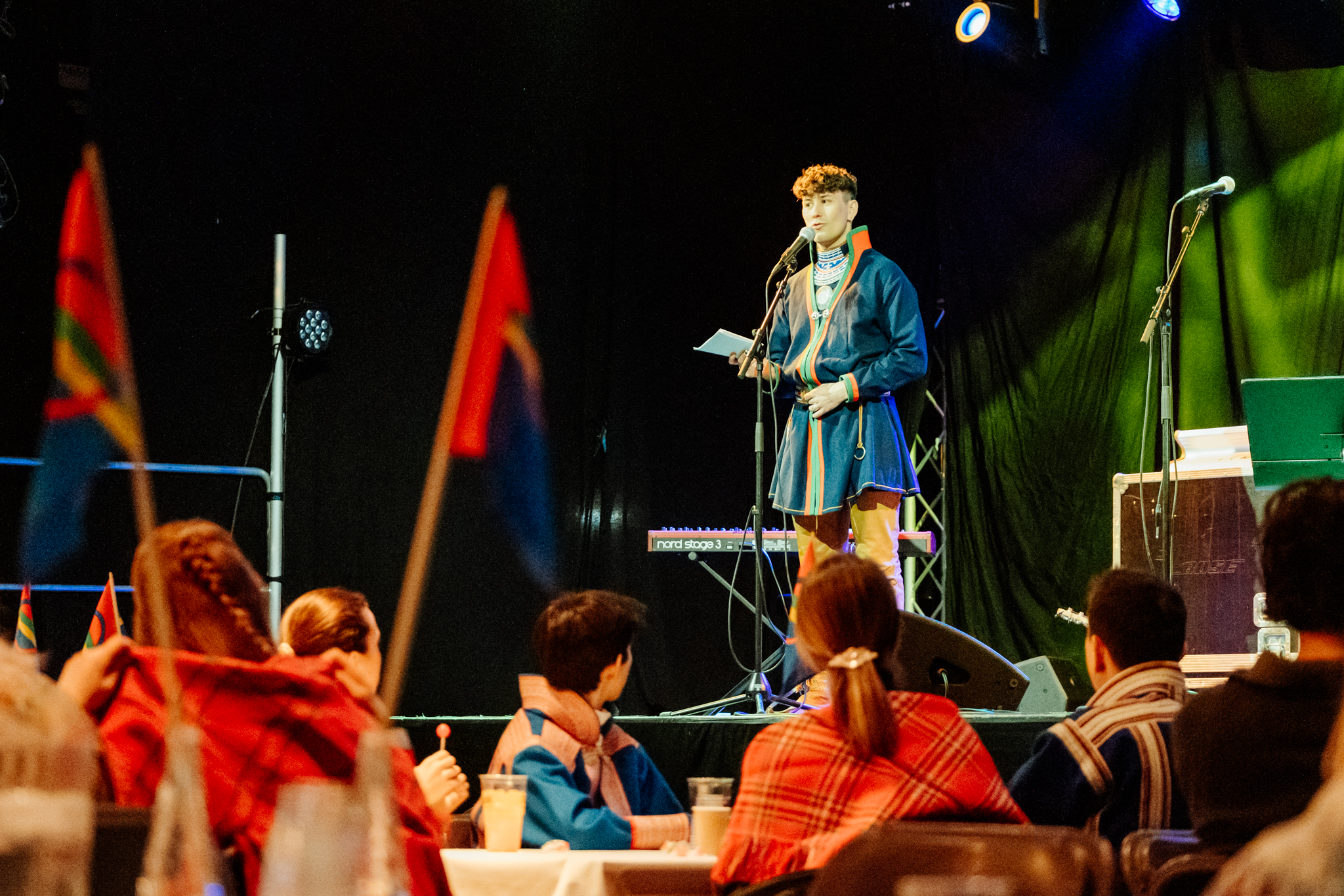 En mann i kofte står på en scene sammen med samiske flagg
