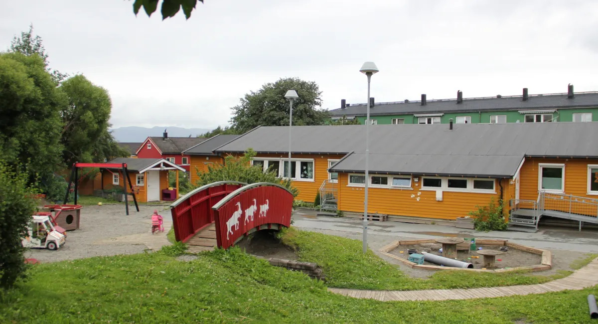 Gult barnehagehus med lekeplass forran og rød bro.