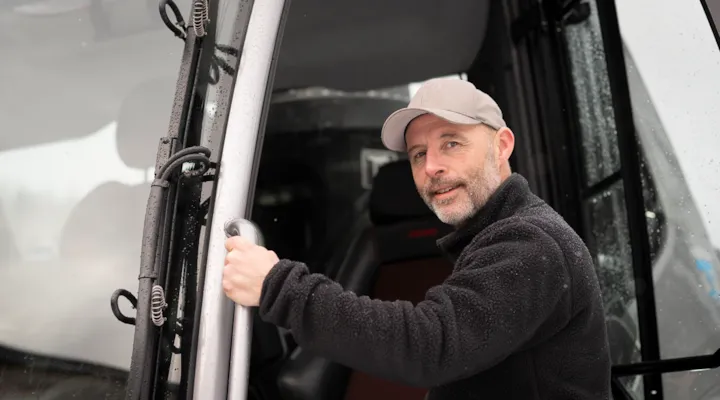 Roger har sorte klær på seg. Står foran løypemaskinen i skogen i Granåsen.