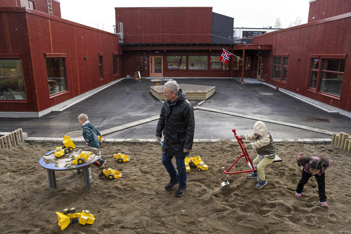 Barn leker, ansatt er sammen med dem, med barnehagen i bakgrunnen.