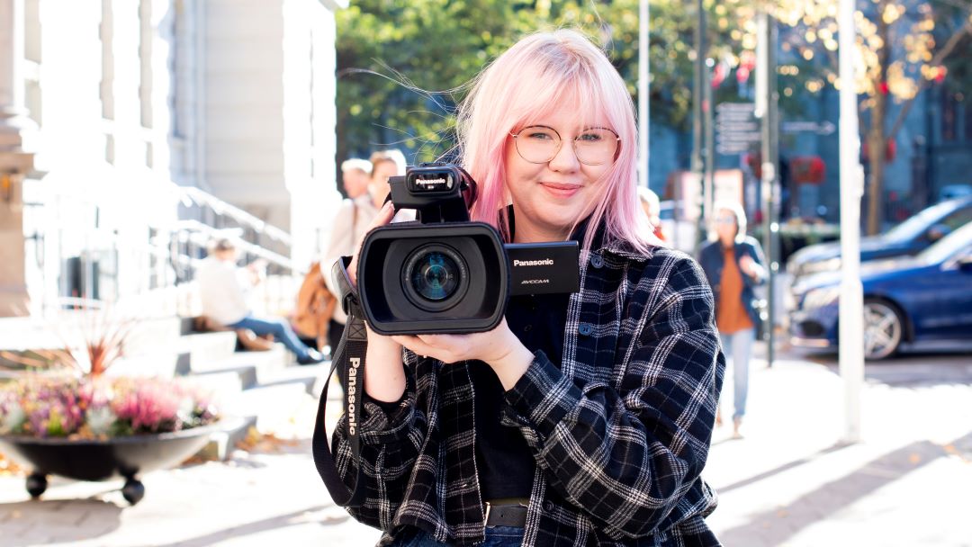 Dame som smiler og holder et videokamera utenfor r&aring;dhuset med nidarosdomen delvis i bakgrunnen