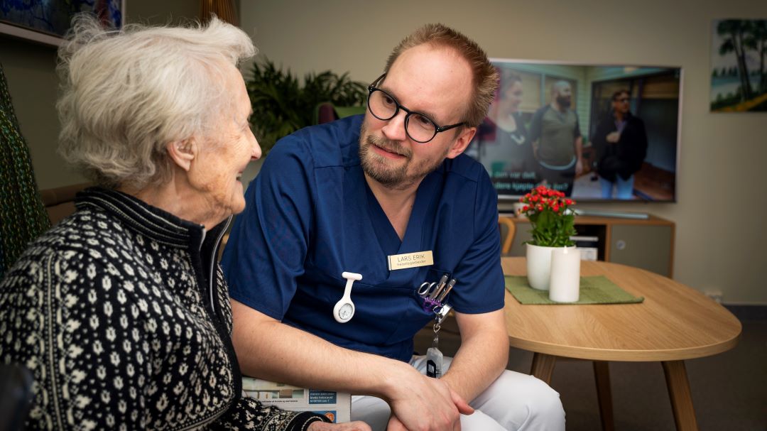 Eldre dame i samtale med mannlig ansatt p&aring; et helse- og velferdssenter. Begge sitter og smiler og ser p&aring; hverandre.