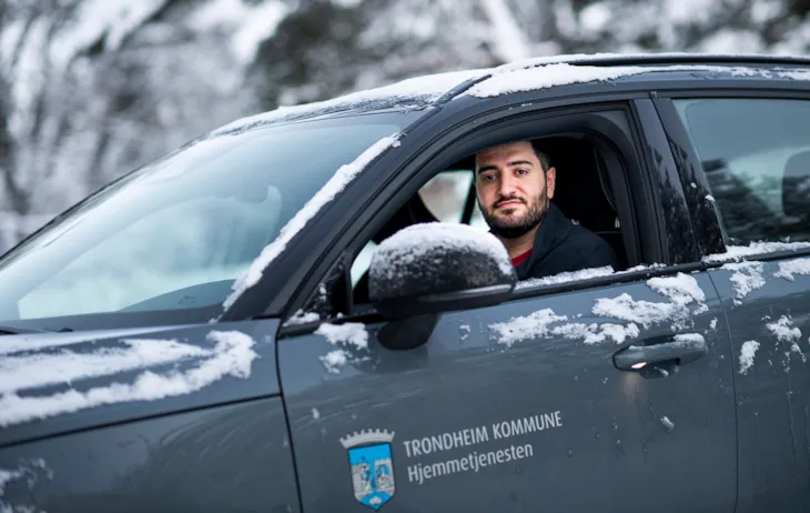 Malik sitter i hjemmetjenestens bil, med nedrullet vindu, og ser i kamera.