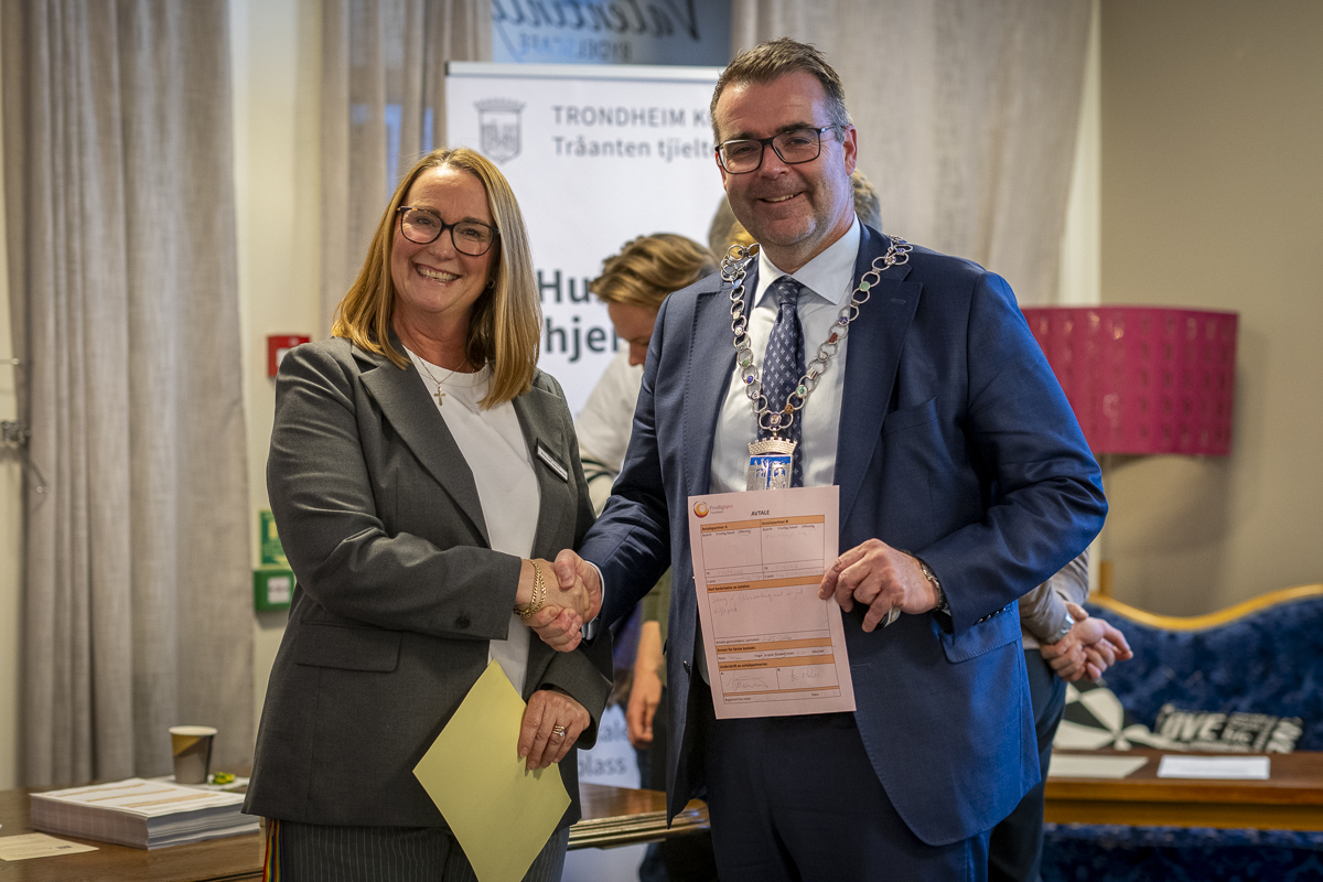 en dame og en mann tar hverandre i hånda, smiler mot kamera og holder frem en kontrakt.