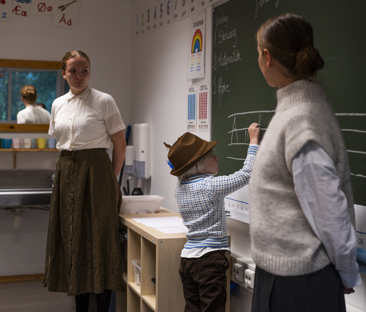 En gutt i 7-&aring;rsalderen skriver bokstaver p&aring; tavla. Han har p&aring; seg hatt og gammeldagse kl&aelig;r, det har ogs&aring; de to l&aelig;rerinnene som st&aring;r ved siden av.