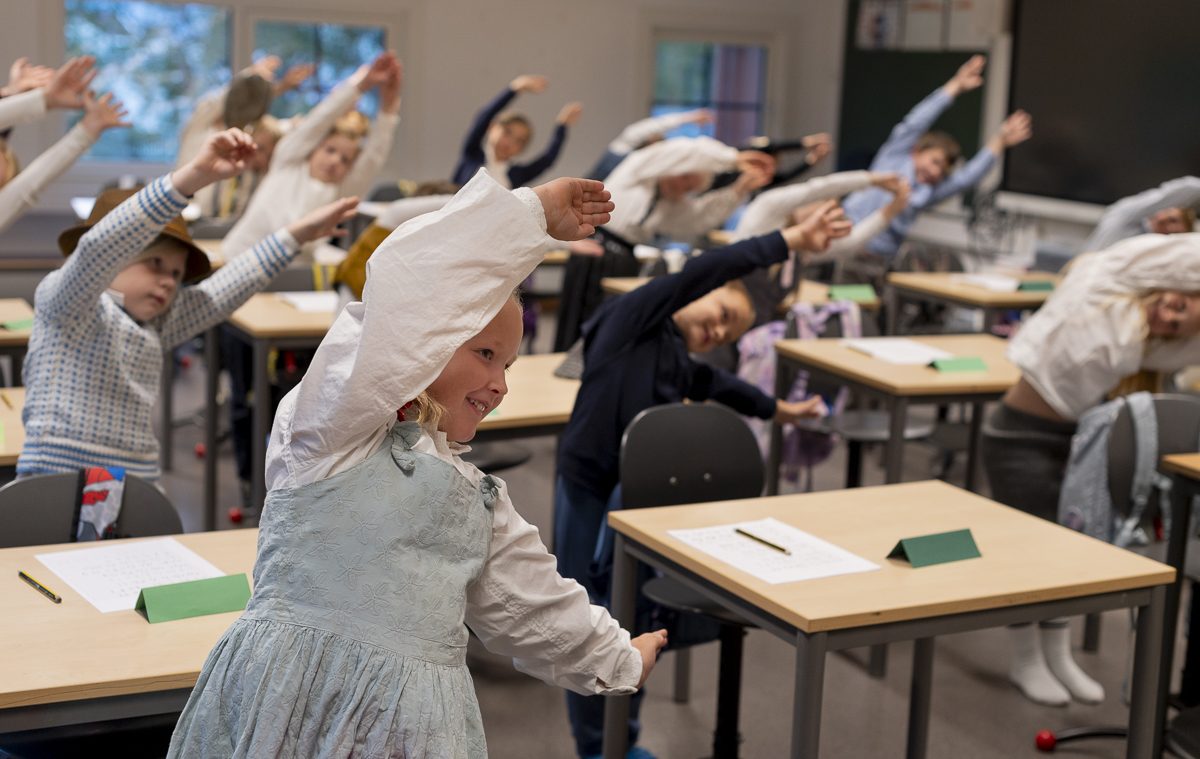 En gruppe elever i sm&aring;skolen st&aring;r bak pultene sine, smiler og b&oslash;yer seg mot h&oslash;yre. Barna har p&aring; seg gammeldagse kl&aelig;r.