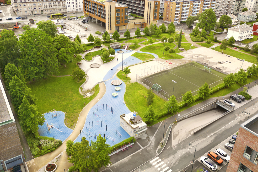  Parkområde med grønne trær, svingete stier og noen personer som går. I bakgrunnen ses flere moderne bygninger.