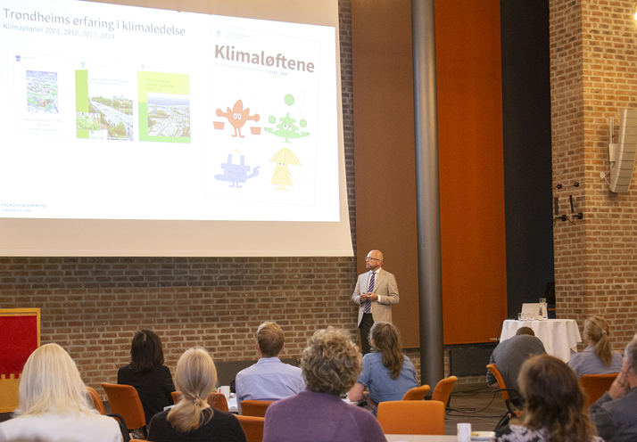 Byrådsleder Kristian Dahlberg Hauge står på scenen og snakker til publikum. På skjermen bak er det en presentasjon som heter Klimaløftene.