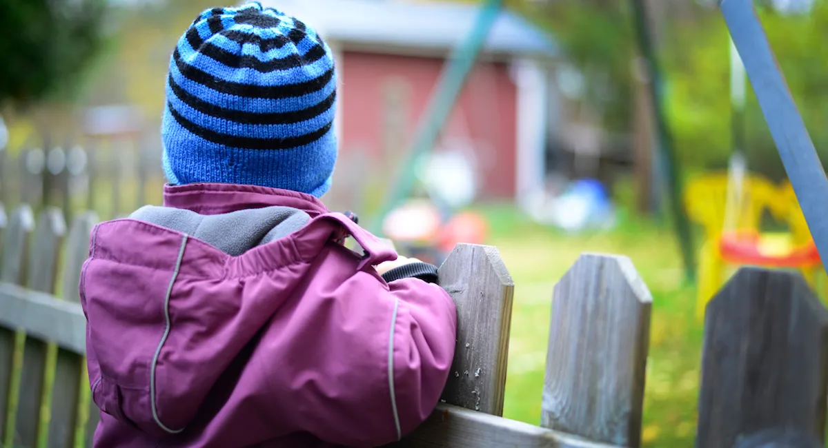 Foto av barn i utedress som står med ryggen til ved et gjerde