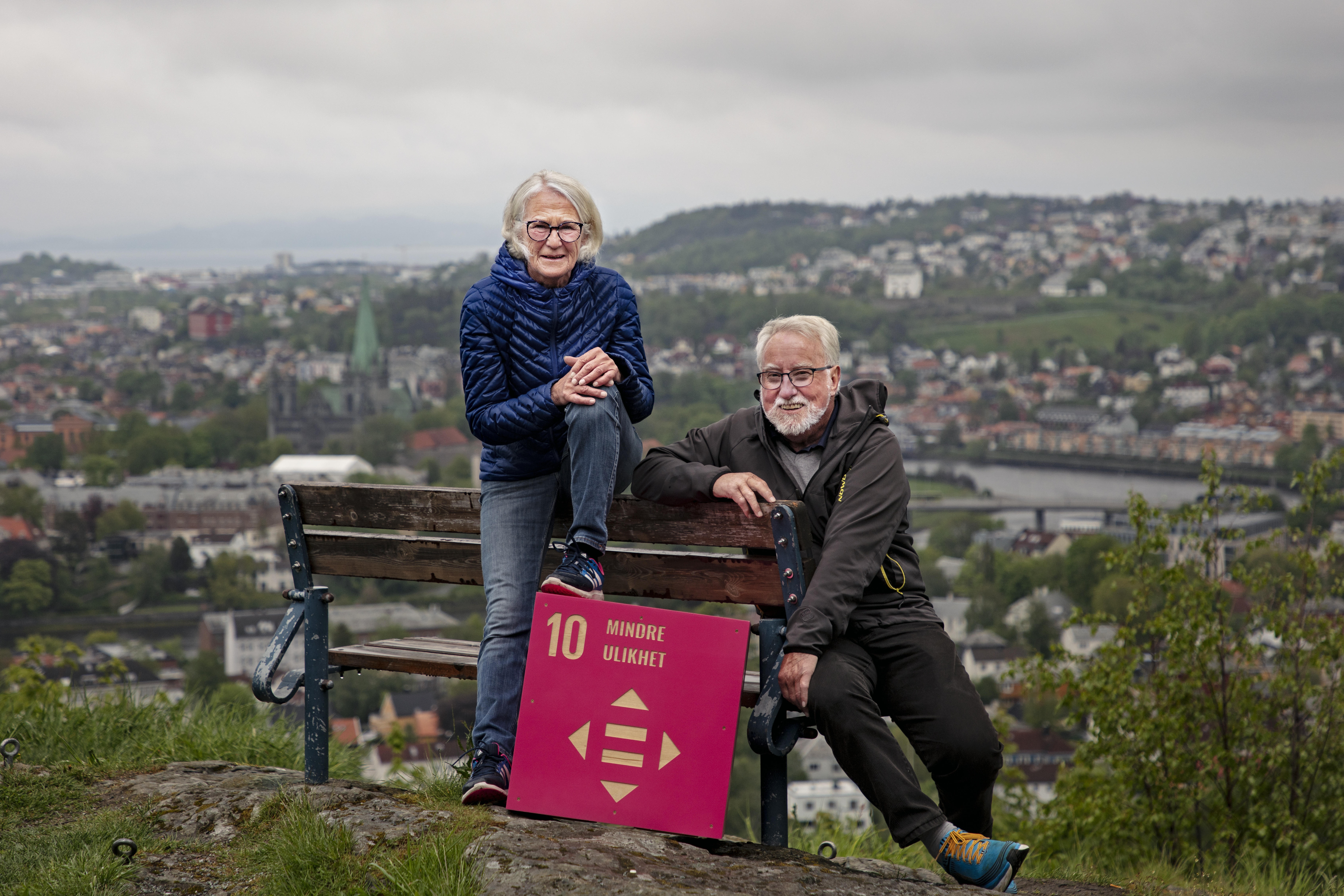 To eldre som sitter på en benk med utsikt til Trondheim i bakgrunnen
