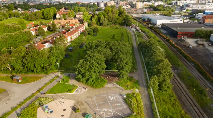 Dronebilde av parken i forgrunnen, omringet av trær, med jernbanelinja på høyre siden. I bakgrunnen er byen, og fjorden.