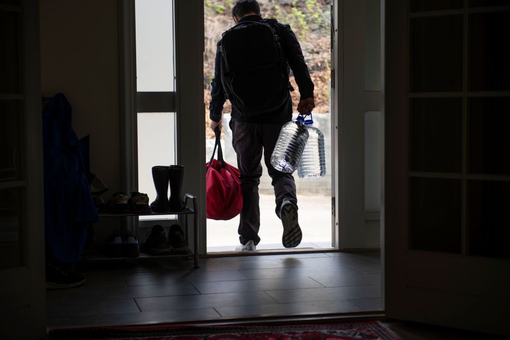 En person sett bakfra b&aelig;rer ryggsekk, en r&oslash;d bag og to store vannkanner ut av ytterd&oslash;ren til et hus