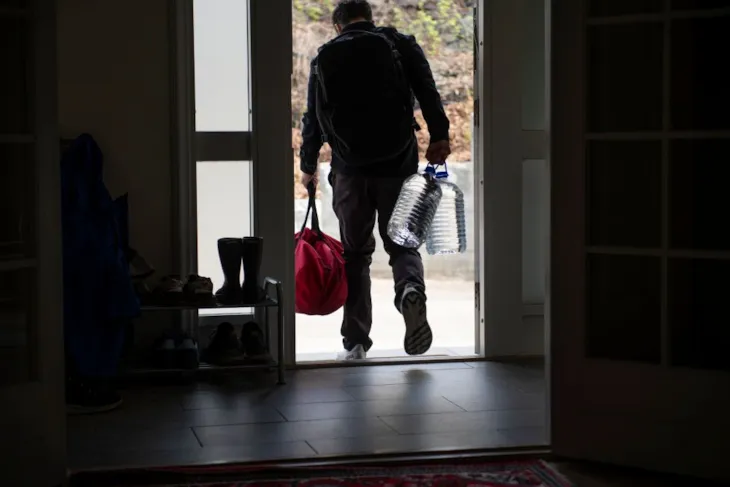 En person sett bakfra bærer ryggsekk, en rød bag og to store vannkanner ut av ytterdøren til et hus