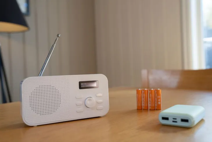 En hvit DAB-radio står på et trebord ved siden av fire oransje batterier og en nødlader (powerbank).