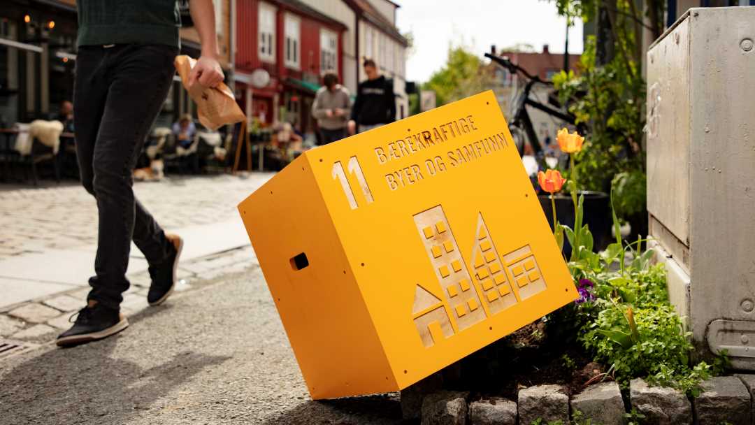 En gul kloss med logoen til bærekraftsmål 11, ligger i et blomsterbed langs en gate på Bakklandet.