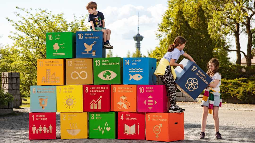 Bilde av de 17  bærekraftsmålene stablet i fire høyder. En gutt sitter oppå, mens to jenter løfter opp det siste bærekraftsmålet.